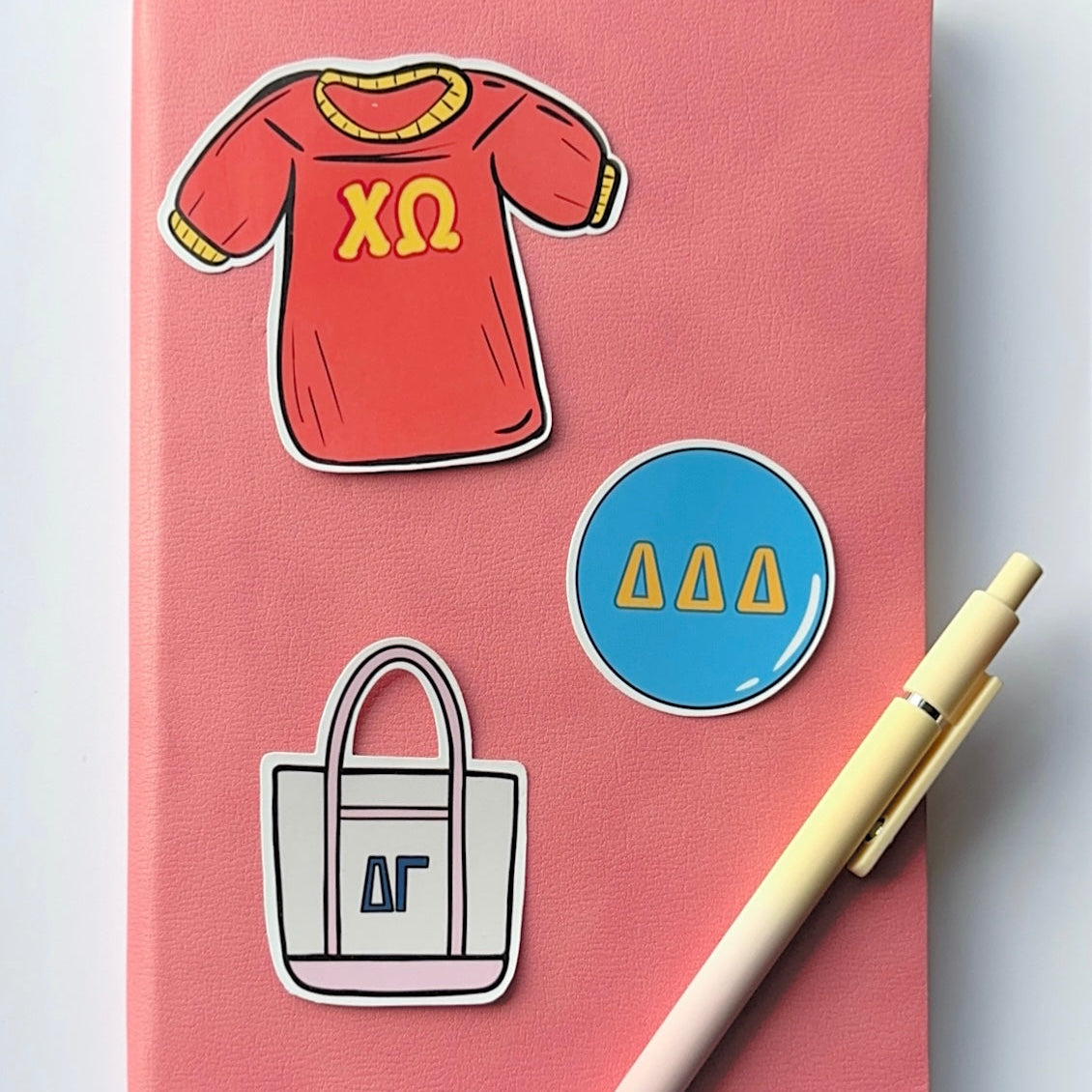 A pink hardcover notebook with decorative stickers, including a red t-shirt with yellow Chi Omega Greek letters, a white tote bag with navy blue Delta Gamma Greek letters, and a blue circle with yellow Delta Delta Delta Greek Letters. A pastel yellow to pink ombre pen rests on the right side, and a pink ribbon bookmark hangs from the bottom left, all on a white background.